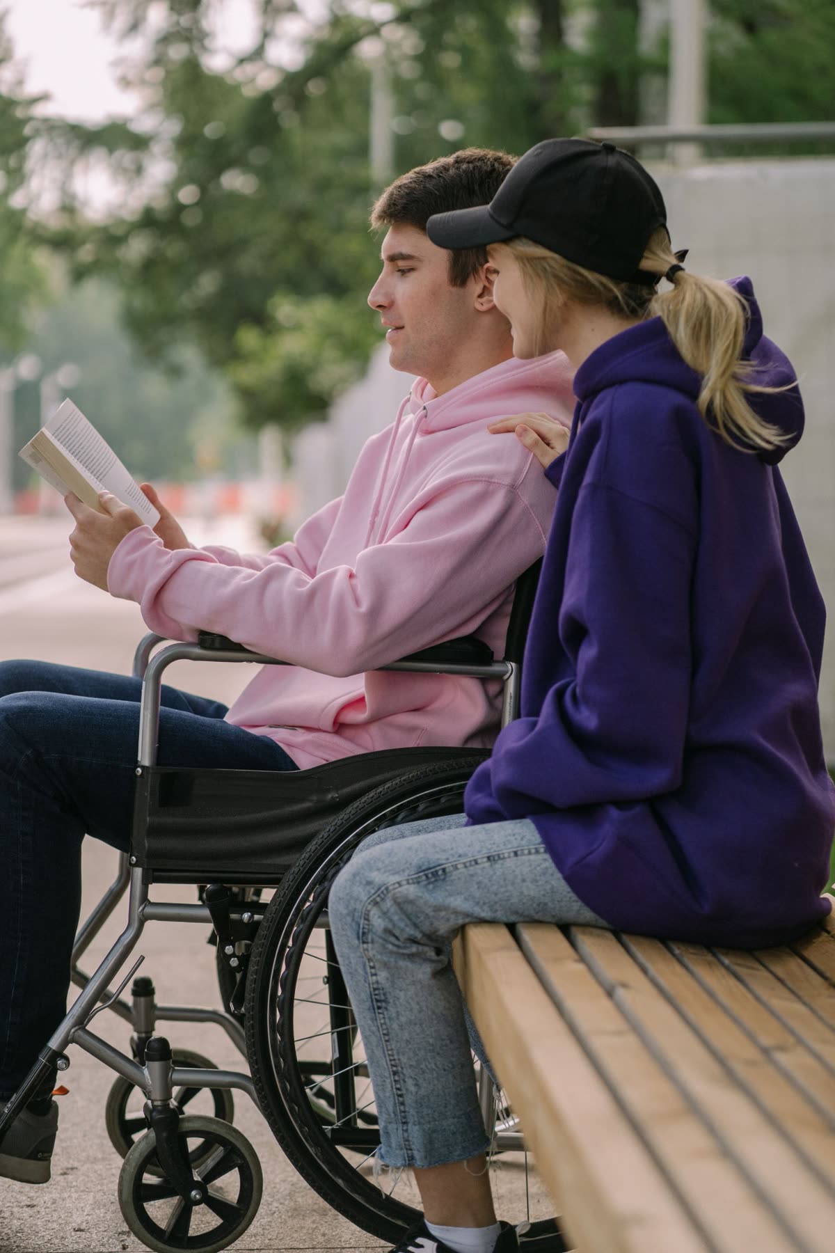 Un jeune homme en fauteuil roulant lit un livre à côté d'une amie assise sur un banc
